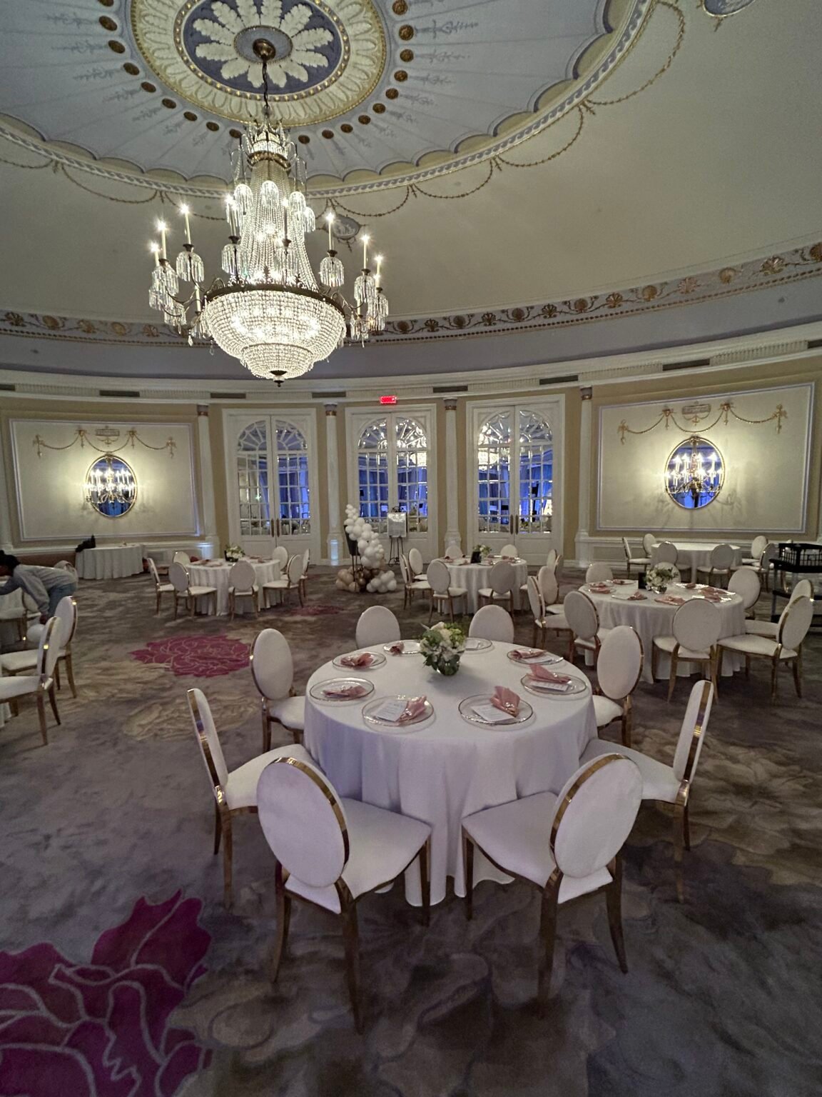 Salle de bal avec lustre de cristal, tables rondes napp&eacute;es de blanc et chaises dos ovale or et blanc dress&eacute;es par &Eacute;venox