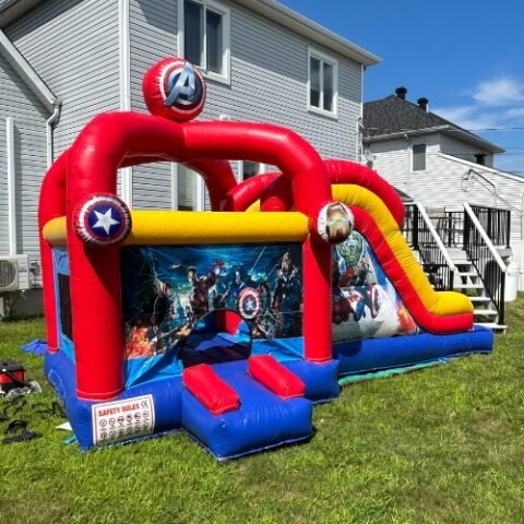 Jeu gonflable Avengers install&eacute; sur une pelouse&nbsp;: arches rouges surmont&eacute;es d'un ballon au logo Avengers, &eacute;cussons de Captain America et d'Iron Man sur les arches, affiche des Avengers imprim&eacute;e au fond et glissade jaune &agrave; droite