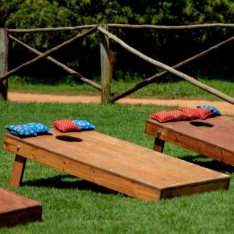 Deux planches de cornhole en bois teint&eacute; fonc&eacute;, inclin&eacute;es sur leurs pieds, pos&eacute;es sur le gazon devant une cl&ocirc;ture de perches&nbsp;; des sacs bleus et rouges &agrave; motif imprim&eacute; sont pos&eacute;s sur les planches