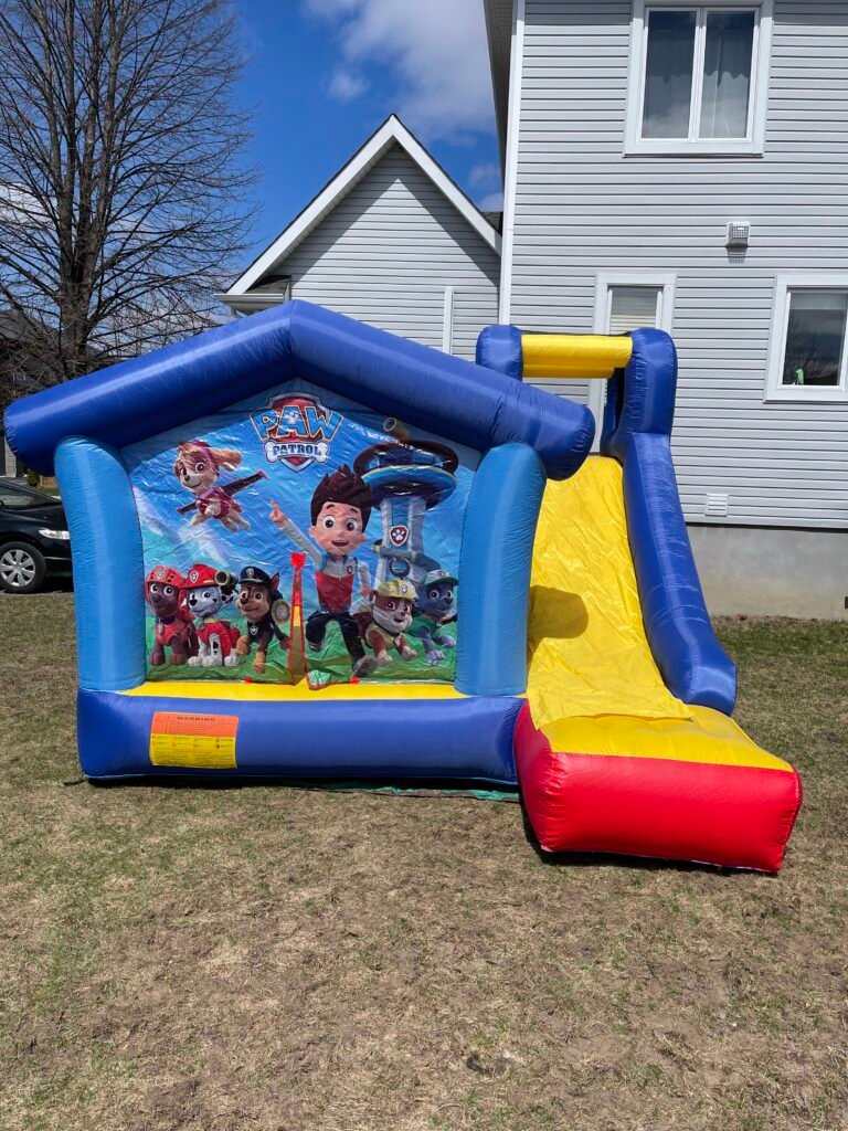 Le jeu gonflable Pat&rsquo; Patrouille gonfl&eacute; sur une pelouse&nbsp;: maisonnette &agrave; toit pointu en boudins bleus, paroi arri&egrave;re imprim&eacute;e aux personnages de la s&eacute;rie avec l'&eacute;cusson PAW PATROL en haut du panneau, aire de saut jaune ouverte &agrave; l'avant, glissade jaune bord&eacute;e de bleu qui sort du c&ocirc;t&eacute; droit et retombe sur un matelas de r&eacute;ception rouge, et autocollant d'avertissement orange et jaune coll&eacute; en bas &agrave; gauche de la base