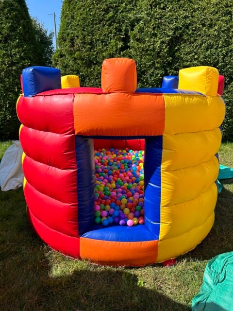 Jeu gonflable Forteresse install&eacute; sur une pelouse&nbsp;: enceinte ronde &agrave; boudins horizontaux rouges, bleus et jaunes, cr&eacute;neaux orange et bleus sur le dessus, ouverture frontale ferm&eacute;e d'un filet, int&eacute;rieur rempli de balles de couleur