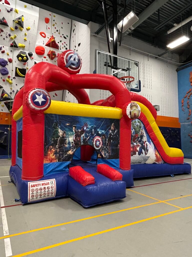 Le jeu gonflable Avengers gonfl&eacute; dans un gymnase&nbsp;: structure rouge, jaune et bleue, ballon Captain America au sommet, entr&eacute;e ouverte au centre, glissade jaune &agrave; droite et panneau « Safety rules » fix&eacute; sur la base