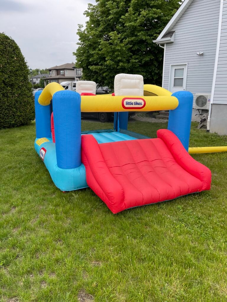 Le jeu gonflable terrain de basket gonfl&eacute; sur une pelouse, vu de trois quarts&nbsp;: base bleu clair, quatre montants bleu fonc&eacute;, portique sup&eacute;rieur jaune portant le logo little tikes, panneau de panier blanc et surface de saut bleue au centre, large glissade rouge descendant vers l'avant droit, parois lat&eacute;rales en filet