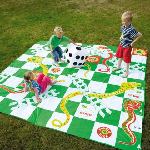 Jeu de serpents et &eacute;chelles g&eacute;ant d&eacute;roul&eacute; sur une pelouse&nbsp;: grande toile &agrave; cases vertes et blanches parcourue de serpents dessin&eacute;s, gros d&eacute; gonflable blanc au centre, trois enfants en train de jouer