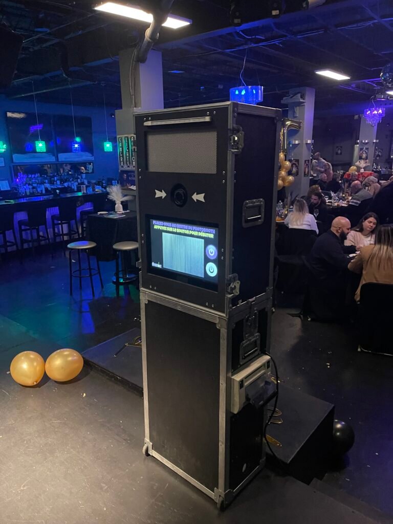 Borne photo &Eacute;venox dans une salle d&rsquo;&eacute;v&eacute;nement en &eacute;clairage bleu, invit&eacute;s attabl&eacute;s en arri&egrave;re-plan et ballons dor&eacute;s au sol