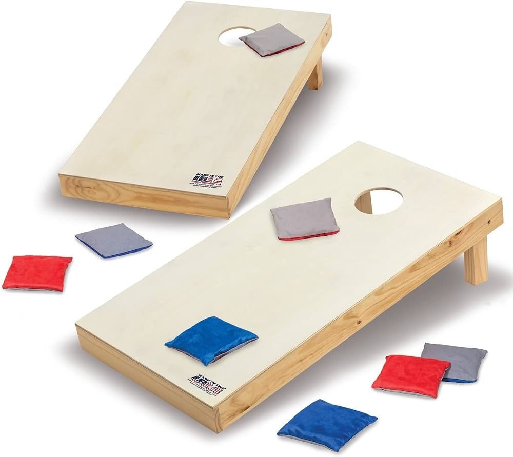 Deux planches de cornhole en bois clair perc&eacute;es d&rsquo;un trou rond, inclin&eacute;es sur leurs pieds sur fond blanc, avec des sacs de lancer rouges, bleus et gris pos&eacute;s dessus et autour