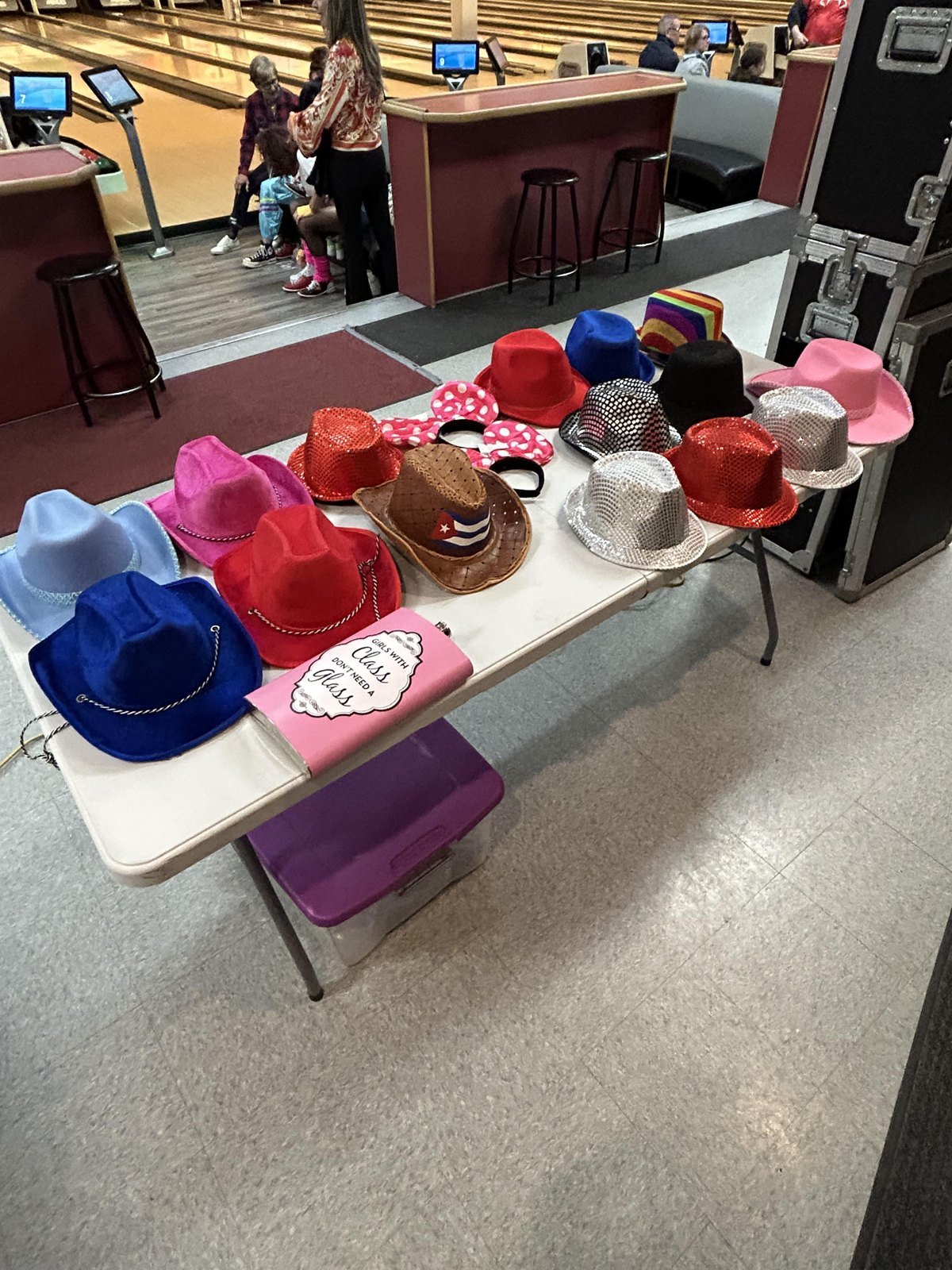 Des chapeaux de fantaisie &mdash; feutres de couleur, panamas &agrave; sequins, oreilles de souris &agrave; pois &mdash; &eacute;tal&eacute;s sur une table pliante blanche dans une salle de quilles, un bac de rangement pos&eacute; dessous et la caisse noire du photobooth &agrave; droite