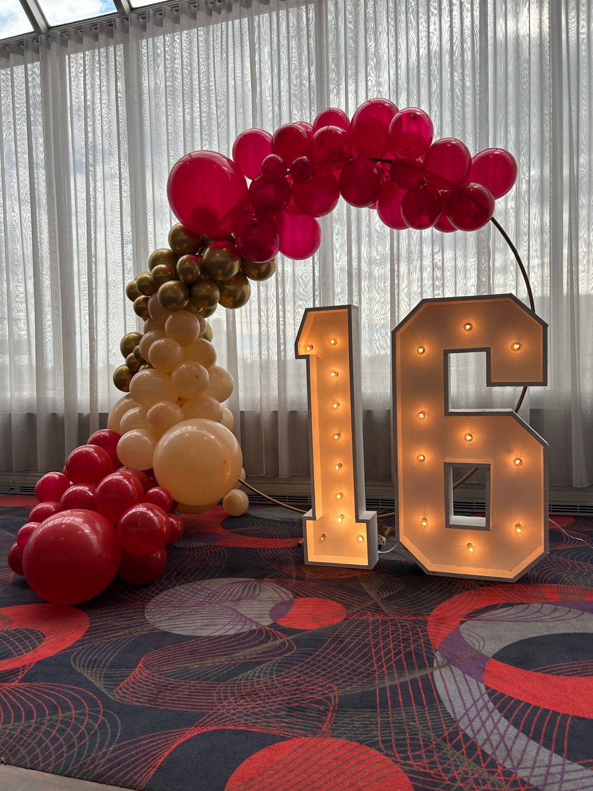 Les chiffres lumineux 1 et 6 devant un rideau blanc, avec une arche de ballons rouges, dor&eacute;s et cr&egrave;me mont&eacute;e &agrave; c&ocirc;t&eacute;