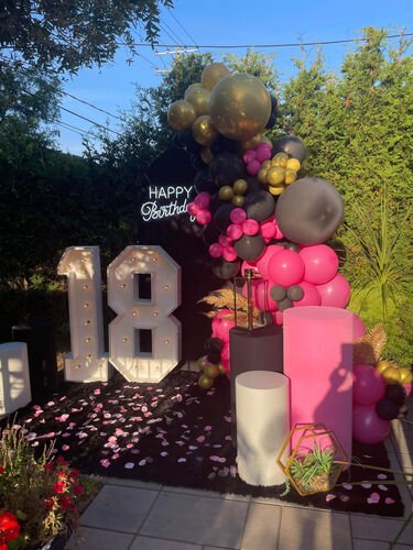 Chiffres lumineux géants formant 18, ampoules allumées, devant une arche de ballons roses, noirs et dorés dans une cour