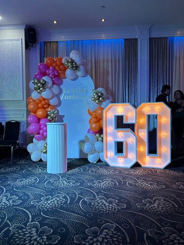 Chiffres lumineux géants formant 60 dans une salle de réception, à côté d'une arche de ballons orange, roses et blancs
