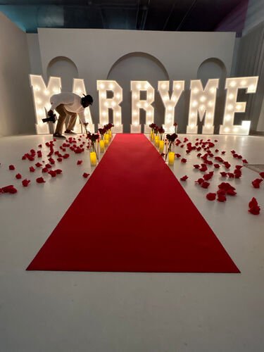 Les grandes lettres lumineuses MARRY ME derri&egrave;re un tapis rouge bord&eacute; de vases, de bougies et de p&eacute;tales