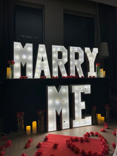 Les lettres lumineuses MARRY ME sur deux rang&eacute;es, avec des roses rouges en vase, des bougies et un c&oelig;ur de p&eacute;tales au sol