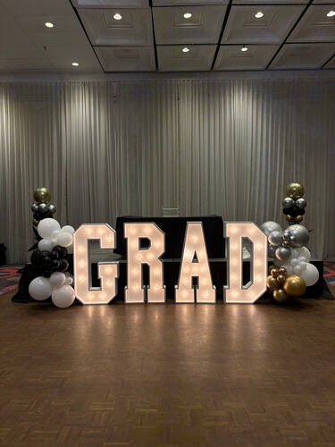 Les lettres lumineuses G R A D align&eacute;es dans une salle de bal, entre deux colonnes de ballons noirs, blancs et dor&eacute;s