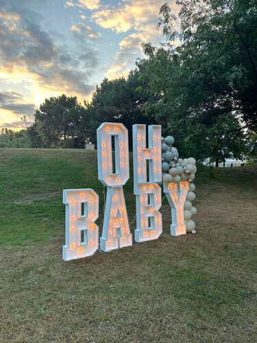 Six lettres lumineuses géantes formant OH BABY sur deux rangs, ampoules allumées, dans un parc au coucher du soleil, avec une colonne de ballons verts à droite