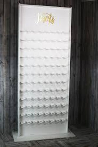 Mur a verres a shooter : panneau de bois blanc portant dix rangees de supports a verres, surmonte du mot Shots en lettres dorees