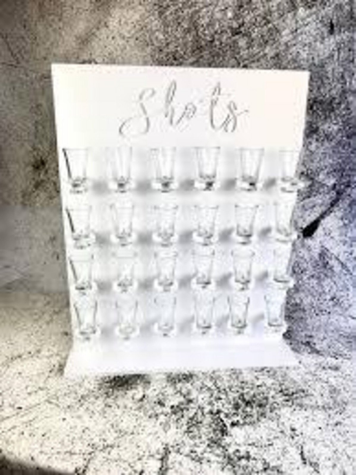 Panneau de bois blanc portant le mot Shots en lettres script, perc&eacute; de rang&eacute;es de trous qui retiennent des verres &agrave; shooter