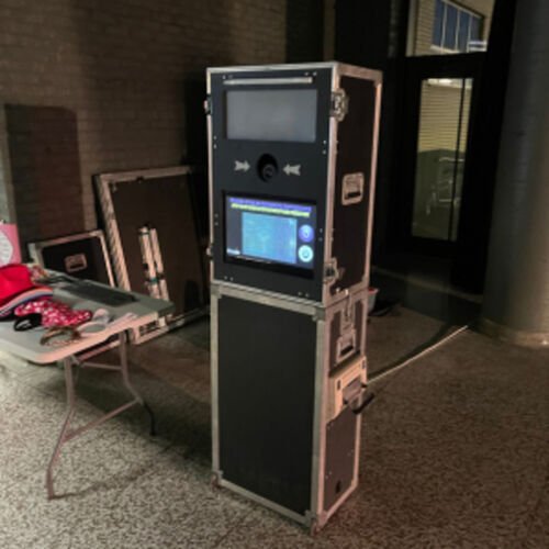 Borne photo noire en caisse de transport, &eacute;cran tactile allum&eacute; et objectif au centre, install&eacute;e dans un hall ; une table d'accessoires est pos&eacute;e &agrave; c&ocirc;t&eacute;